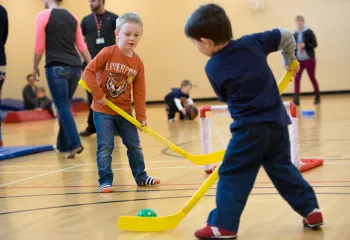 young boys playing floor hockey in gymnasium with family in background