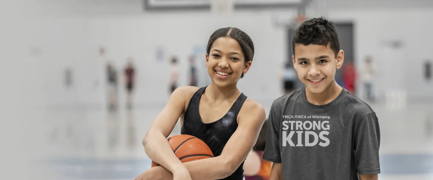 [Image] A girl and boy stand in a gymnasium. The girl holds a basketball. Wordmark in the corners says YMCA-YWCA of Winnipeg Strong Kids