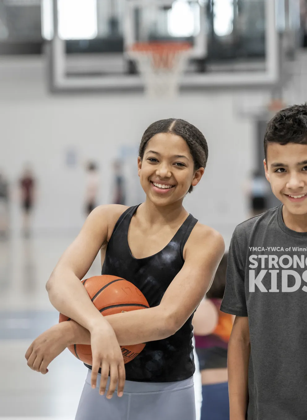 [Image] A girl and boy stand in a gymnasium. The girl holds a basketball. Wordmark in the corners says YMCA-YWCA of Winnipeg Strong Kids