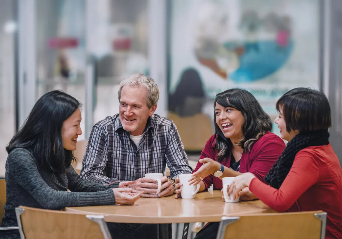 Image of three women and a man sitting at a table laughing with coffee mugs in their hands