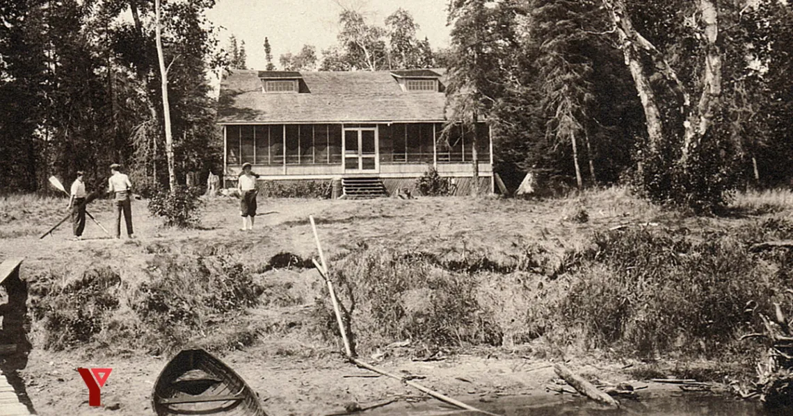 Black and white image of cabin on the shore with three people, paddles and a canoe.