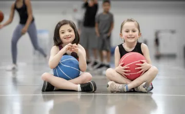 Two young girls sitting with basketballs smiling at camera